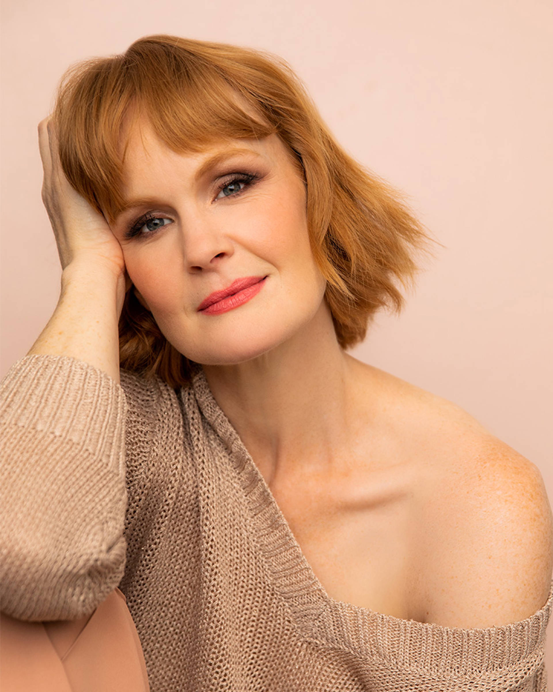 Headshot of a caucasian woman with red medium length hair, dark eyes and red lipstick against a pink background.