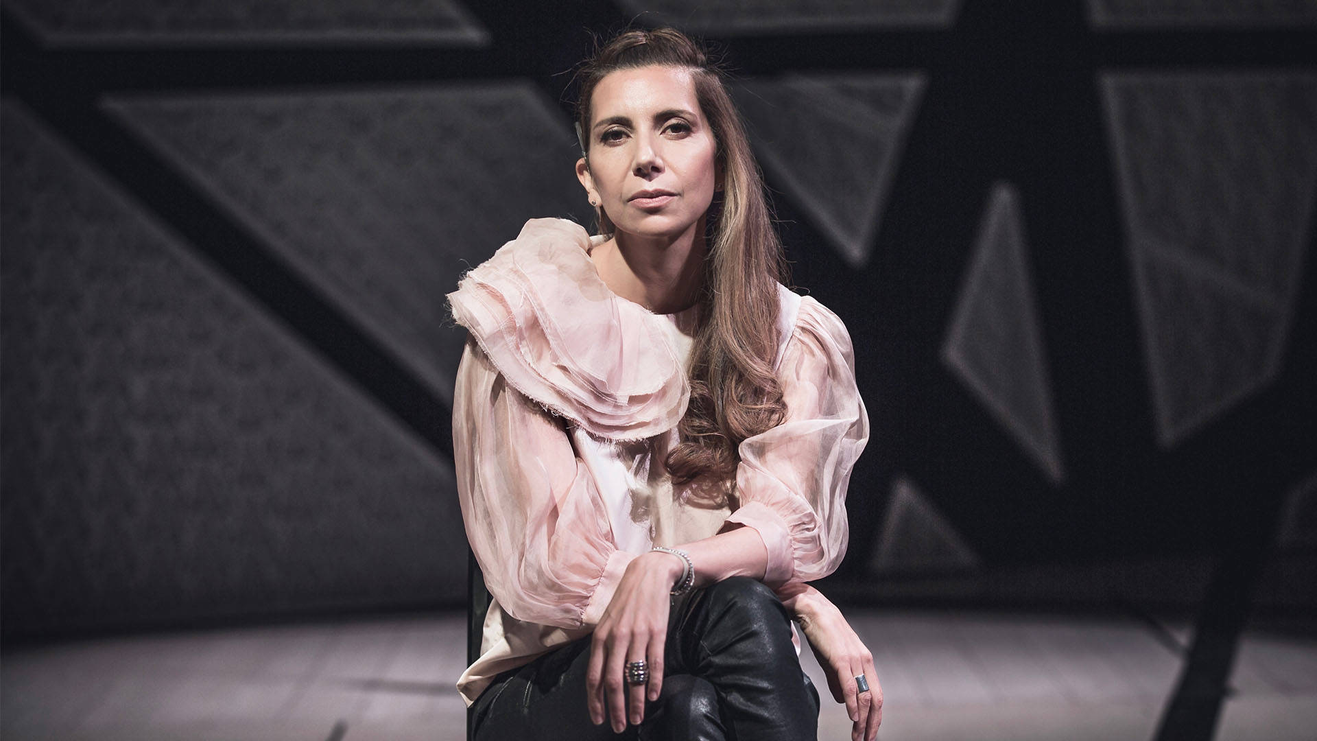 Magos Herrera, a Mexican woman with light brown long hair sits in front of a grey background. She is wearing a light pink blouse and black leather pants with her legs crossed.