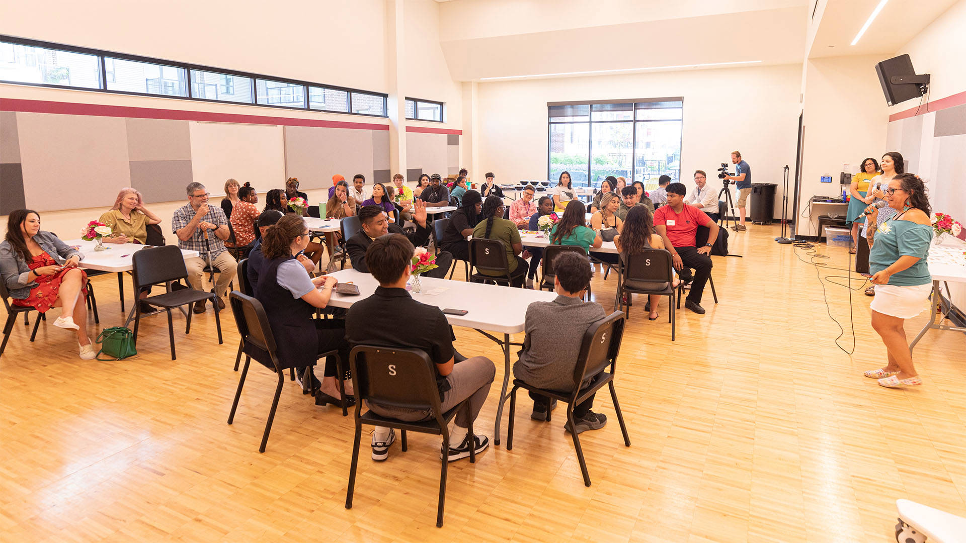 Students sitting at tables in a large room with a wooden floor. A woman on the right speaks into a microphone.