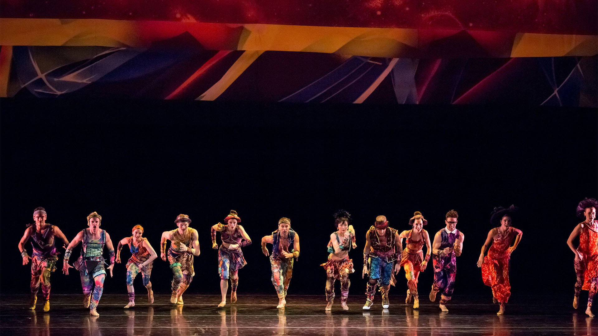 An ensemble of dancers in multicolored random clothing running towards the edge of the stage in a horizontal line.