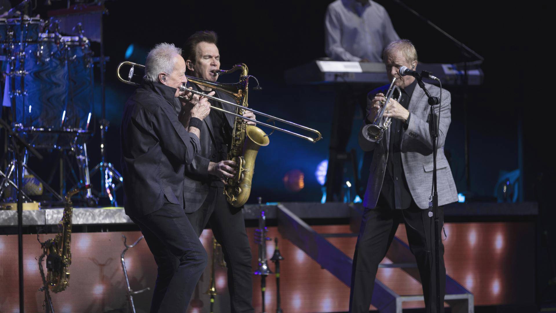 Three men on stage playing trombone, saxophone and trumpet.