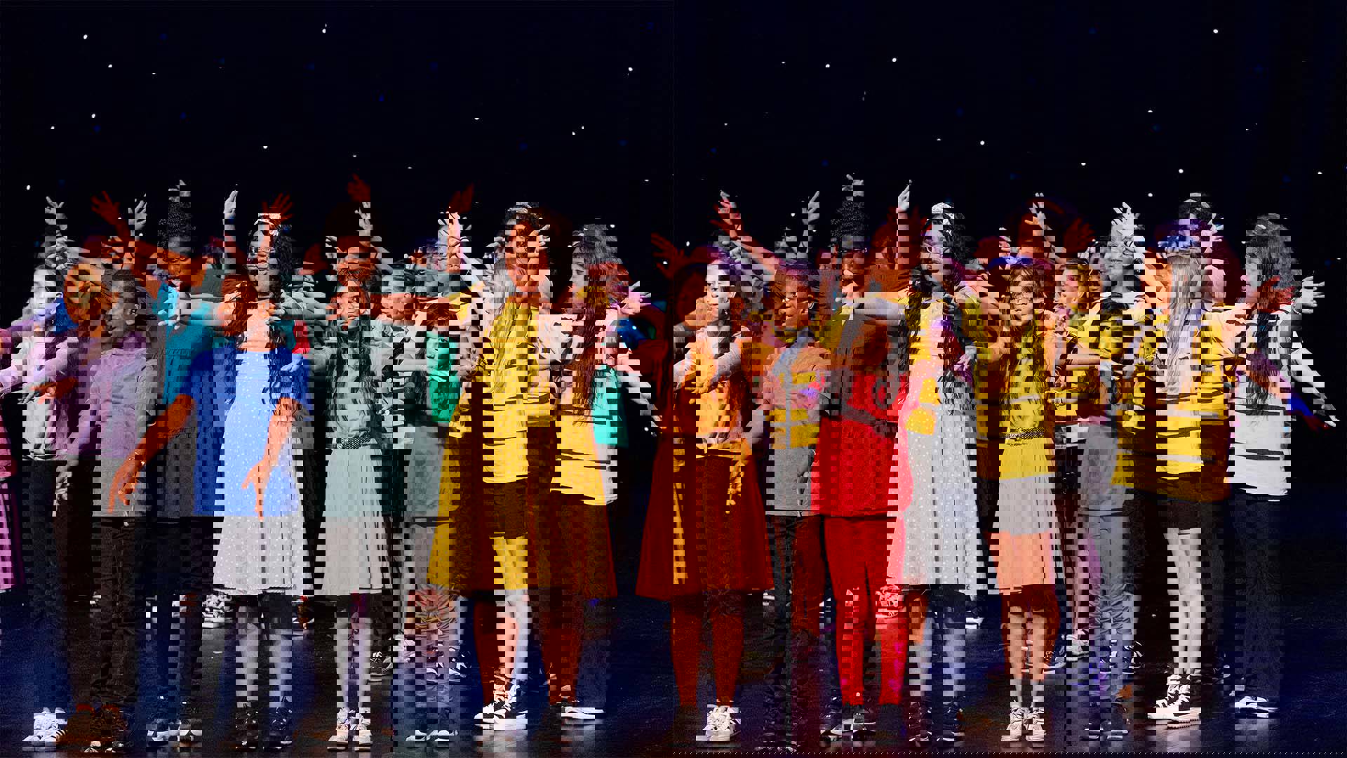 Young students in primary color outfits performing on a dark stage. 