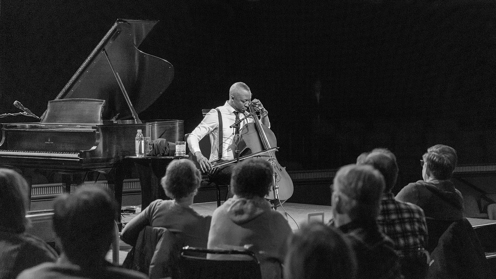 An African American man with a short mohawk and beard sitting and singing into a microphone. He is holding a cello in playing position and there is a grand piano behind him.