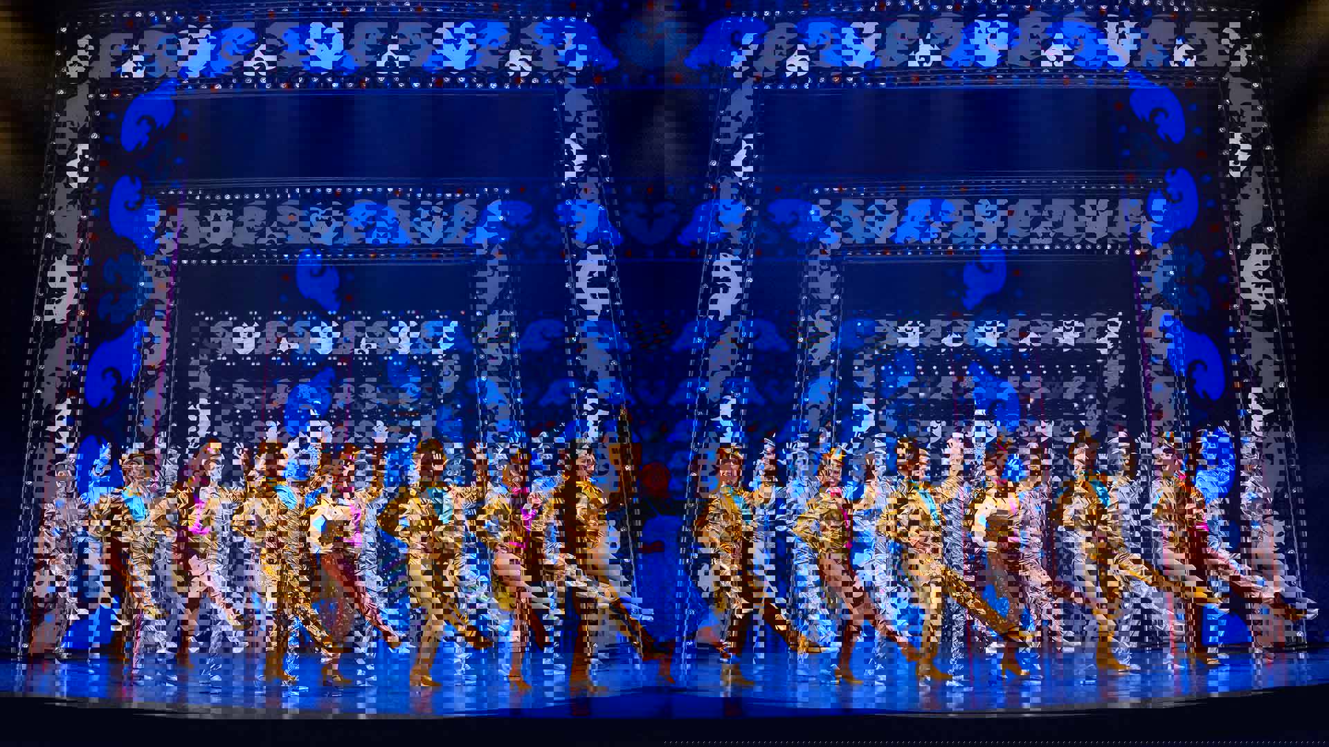 A large group of performers in bright gold attire perform a kick line in front of a blue frame backdrop.