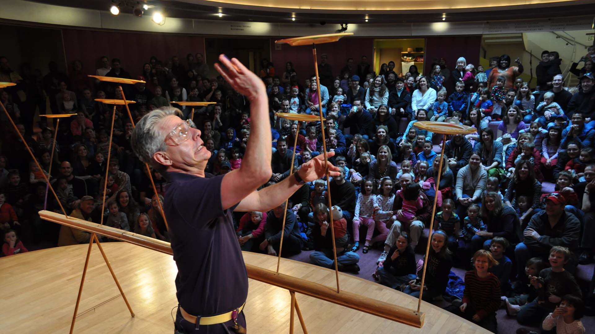 A white man standing on a stage surrounded by an audience of young kids and adults. He is spinning many plates on poles and looking frantic.