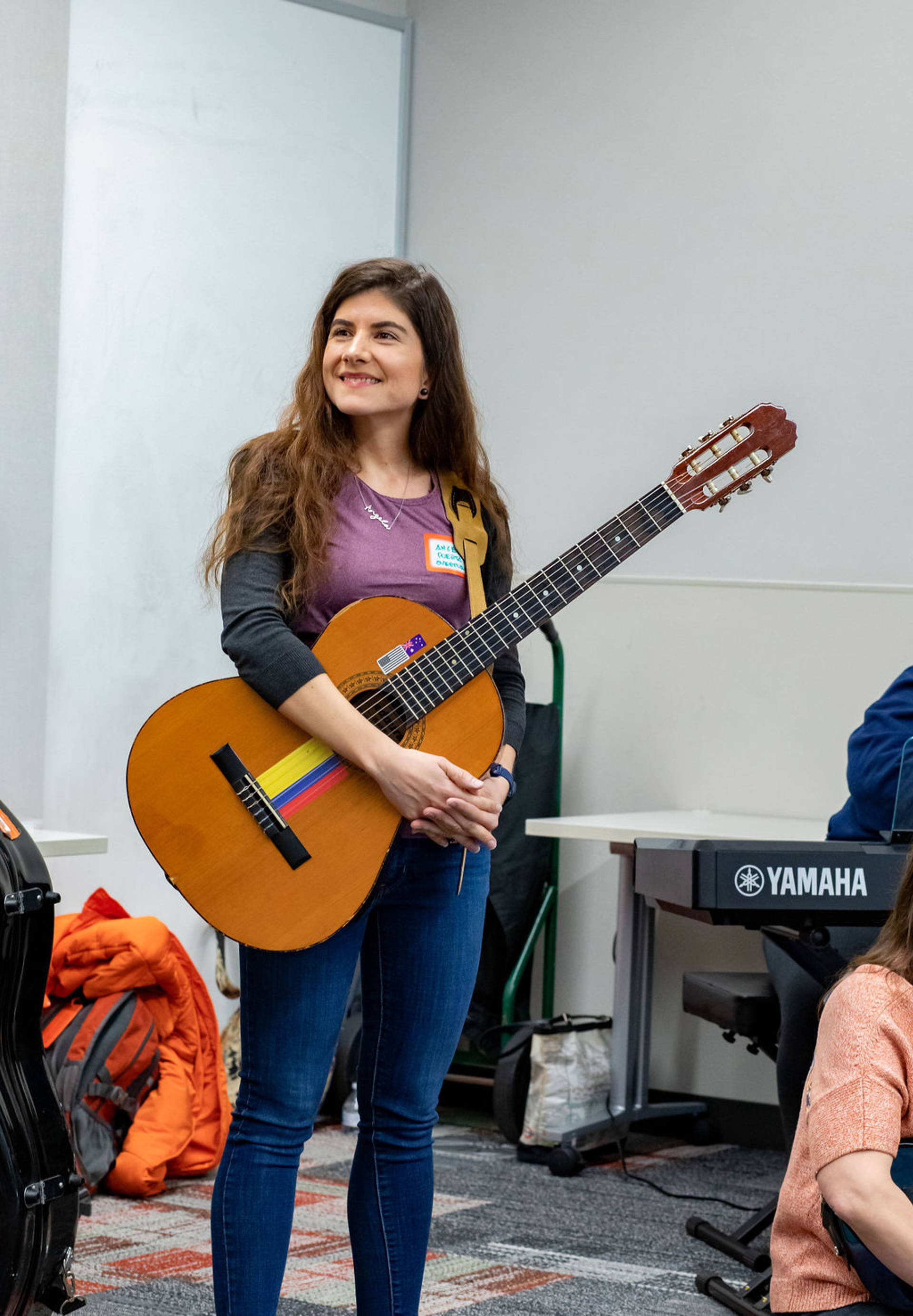 A young woman with medium complexion and long brown hair stands smiling and holding a classical guitar.