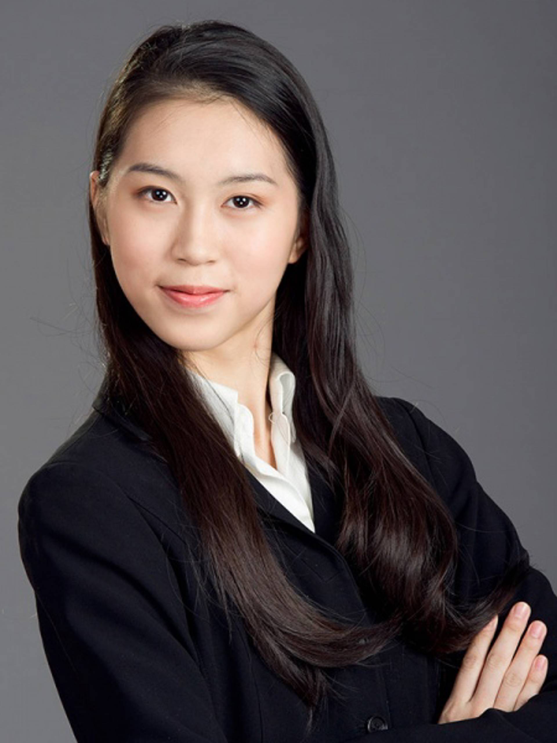 Headshot of a young woman with medium complexion, long black hair, a white collared shirt and a dark jacket against a grey background