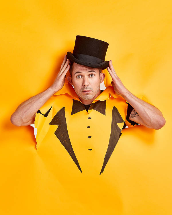 Justin Willman, a caucasian man with short hair and brown eyes. His head and arms have broken through an orange wall. Tuxeo lapels and a bowtie are drawn on the wall where his chest would be. His hands are propping a tophat on his head.