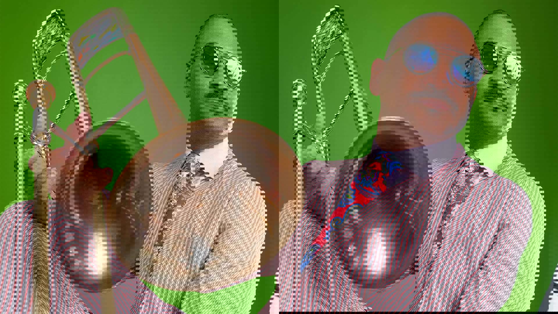 A Black man with buzzed hair, a cropped mustache and sunglasses wearing a red pin striped suit with a red paisley tie, sitting and holding a trombone against his crossed knee.