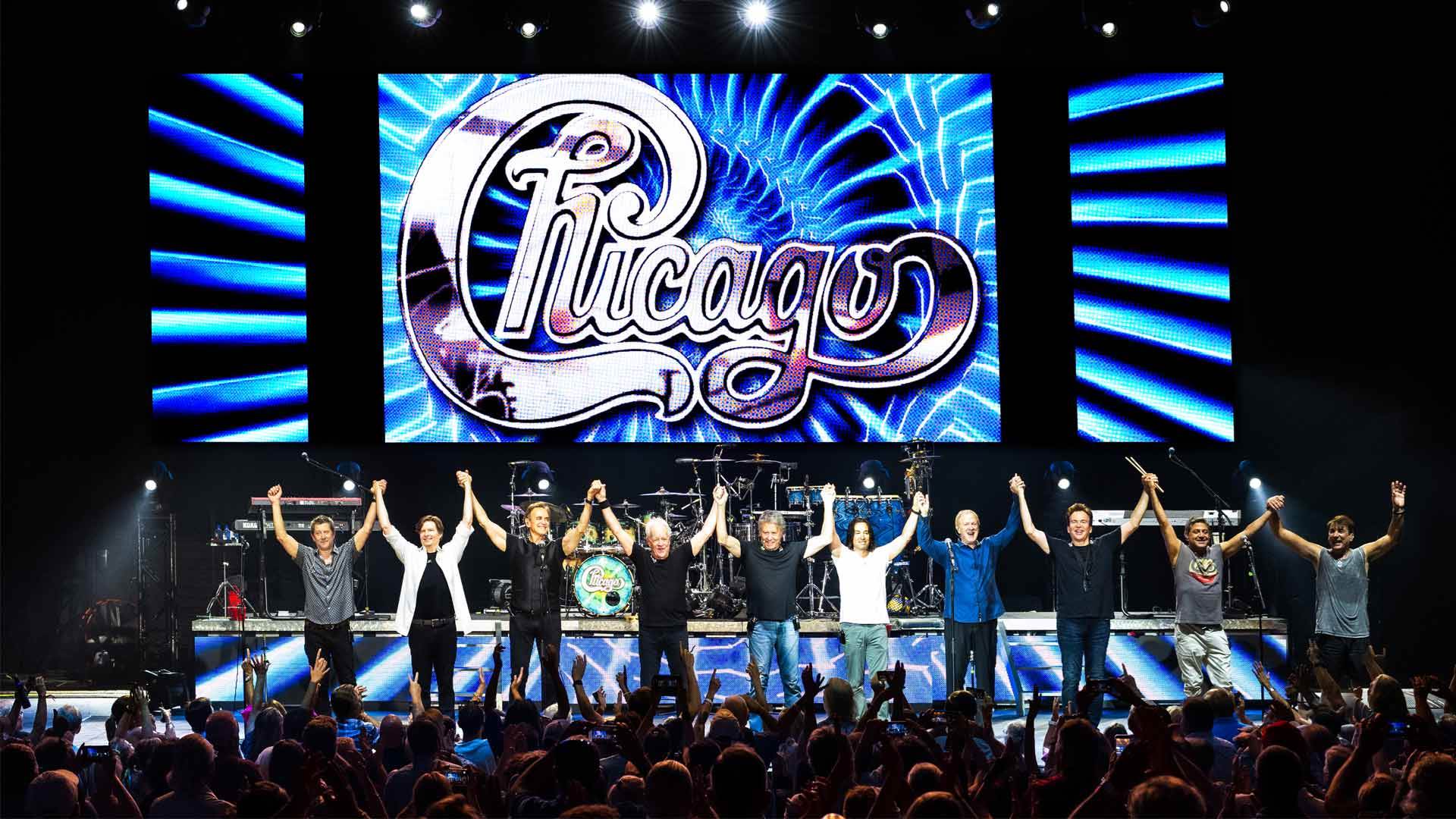Ten people holding each other's hands above their heads in a line on stage in front of a large audience. Behind them is a large screen that says "Chicago".