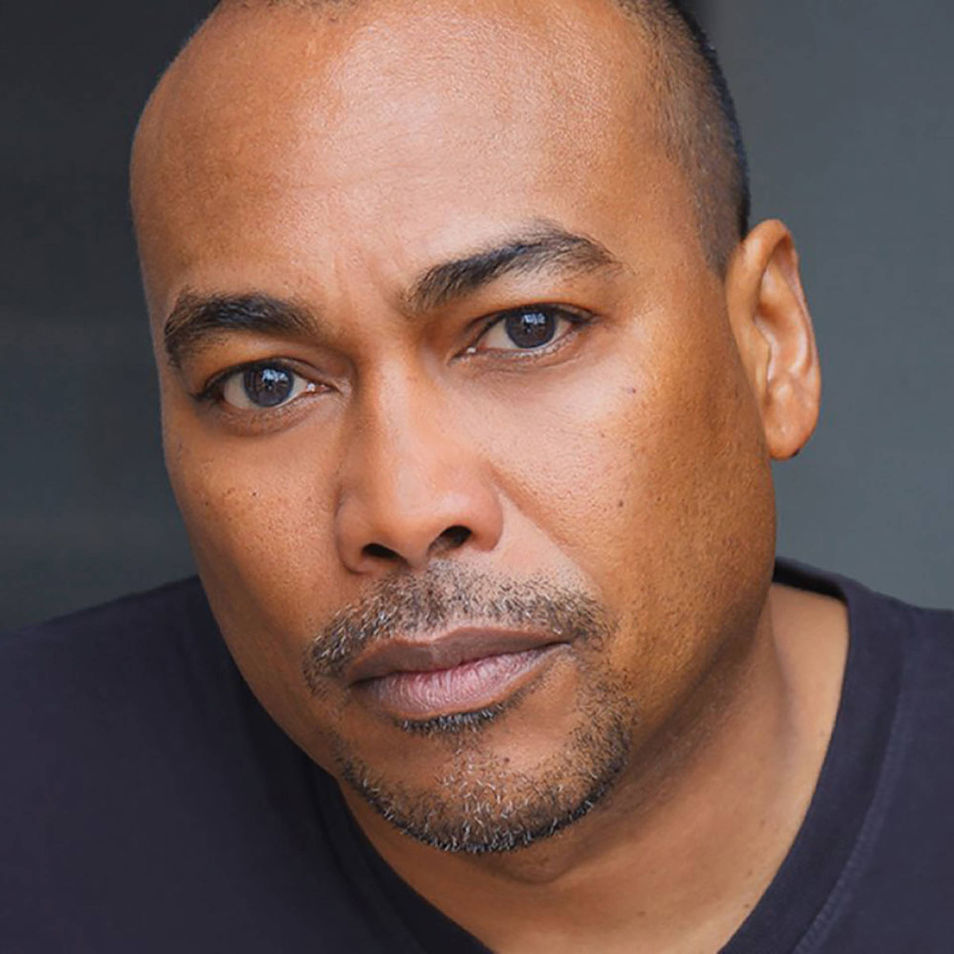 Headshot of an african american man with a bald head, brown eyes and a short goatee in a black shirt.