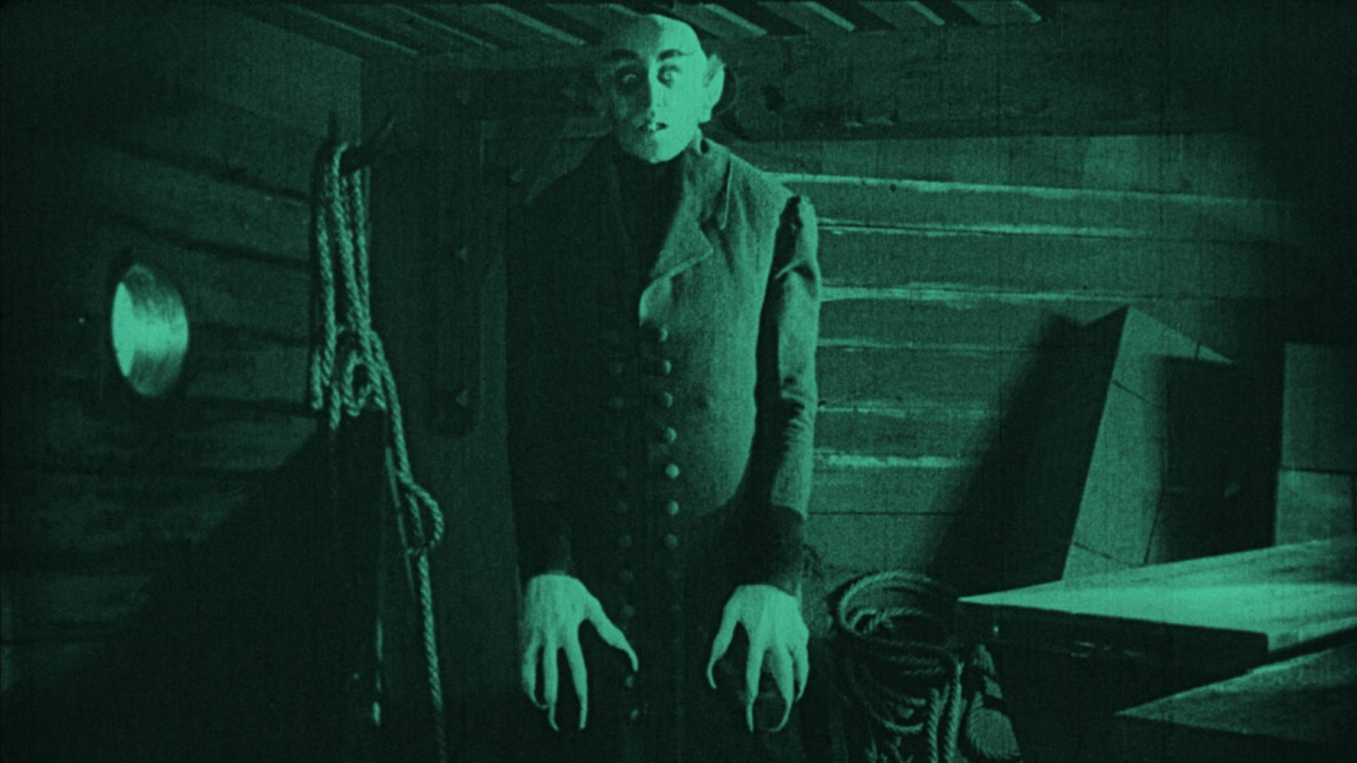 Vintage monotone image of a tall scary figure with long fingers, a bald head, and a dark outfit on standing erect with arms tight to his side and staring at the viewer