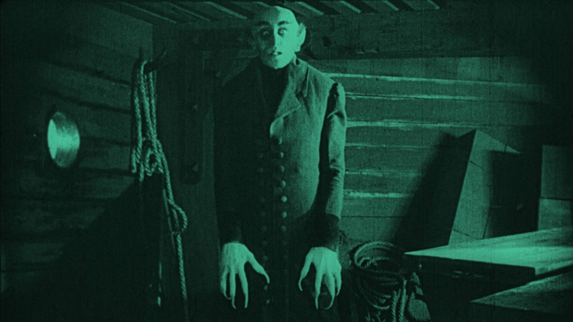 Vintage monotone image of a tall scary figure with long fingers, a bald head, and a dark outfit on standing erect with arms tight to his side and staring at the viewer