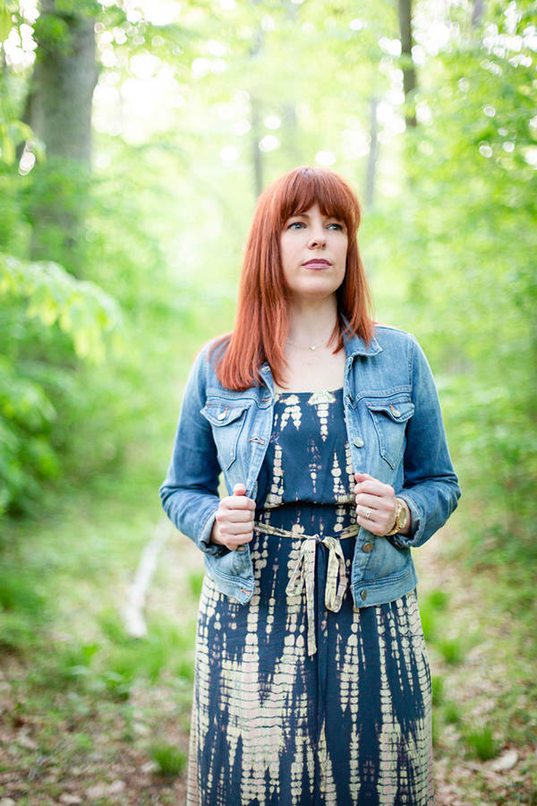 A white woman with reddish-brown, shoulder-length hair. She standing against a blurry wooded background, wearing a jean jacket and a blue dress.