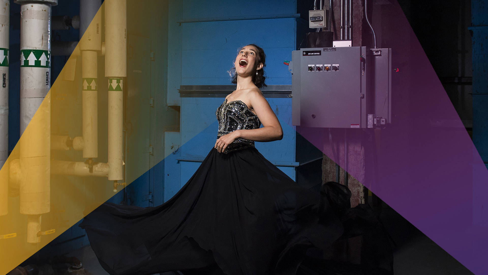 A caucasian woman with medium-length brown hair wearing a strapless black and silver ballgown. She is singing with her head tilted upwards, mouth open, in a mechanical utilities room.