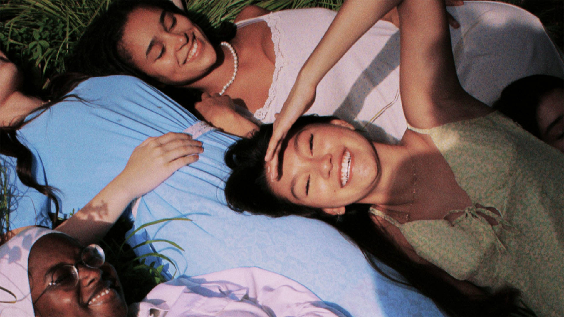 Young teenage girls with dark to medium skin complexions lay in the grass smiling. They are wearing pastel colored clothing.