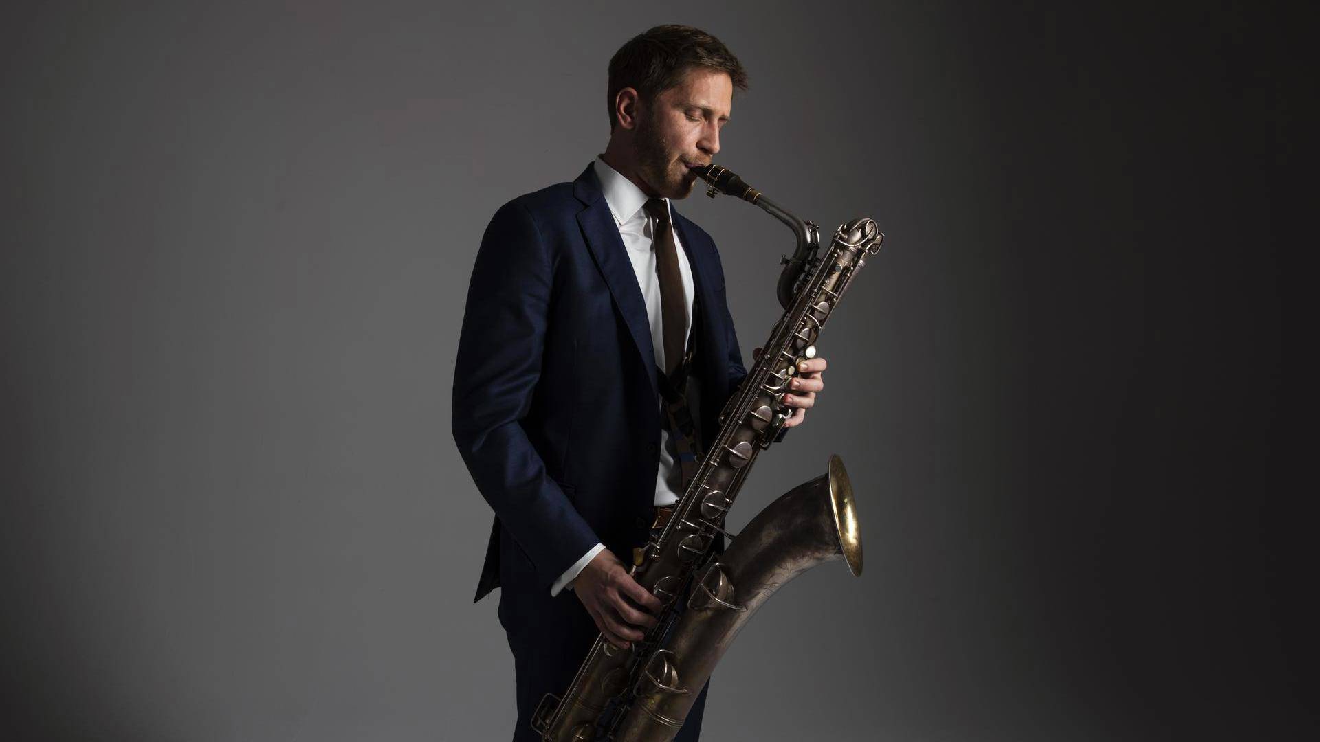 A man with light complexion and dark hair in a black suit playing bari suit. The background is grey.
