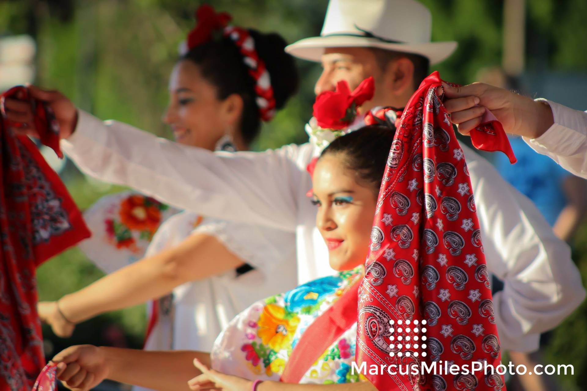 A close up of women and men dancing in white outfits with colorful floral patterns. They are holding read handkerchiefs out in front of them.