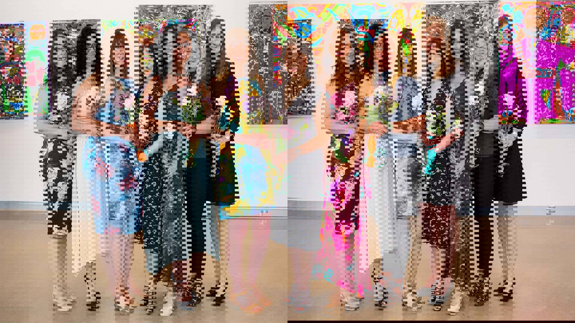 Six women in dresses standing in a.line holding flowers and smiling. A gallery wall is behind them.