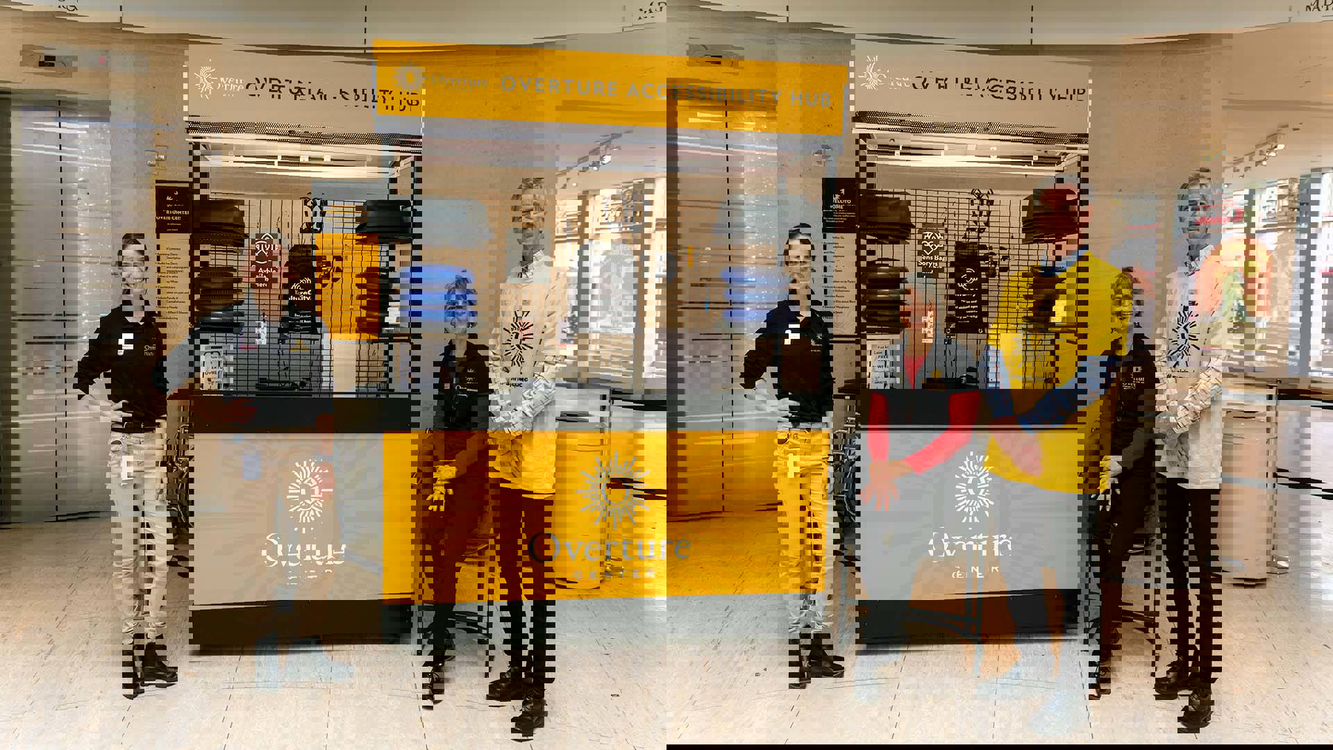 Three Overture staff members with light complexion standing and smiling next to a large metal kiosk with yellow signage that reads "Overture Accessibility Hub."