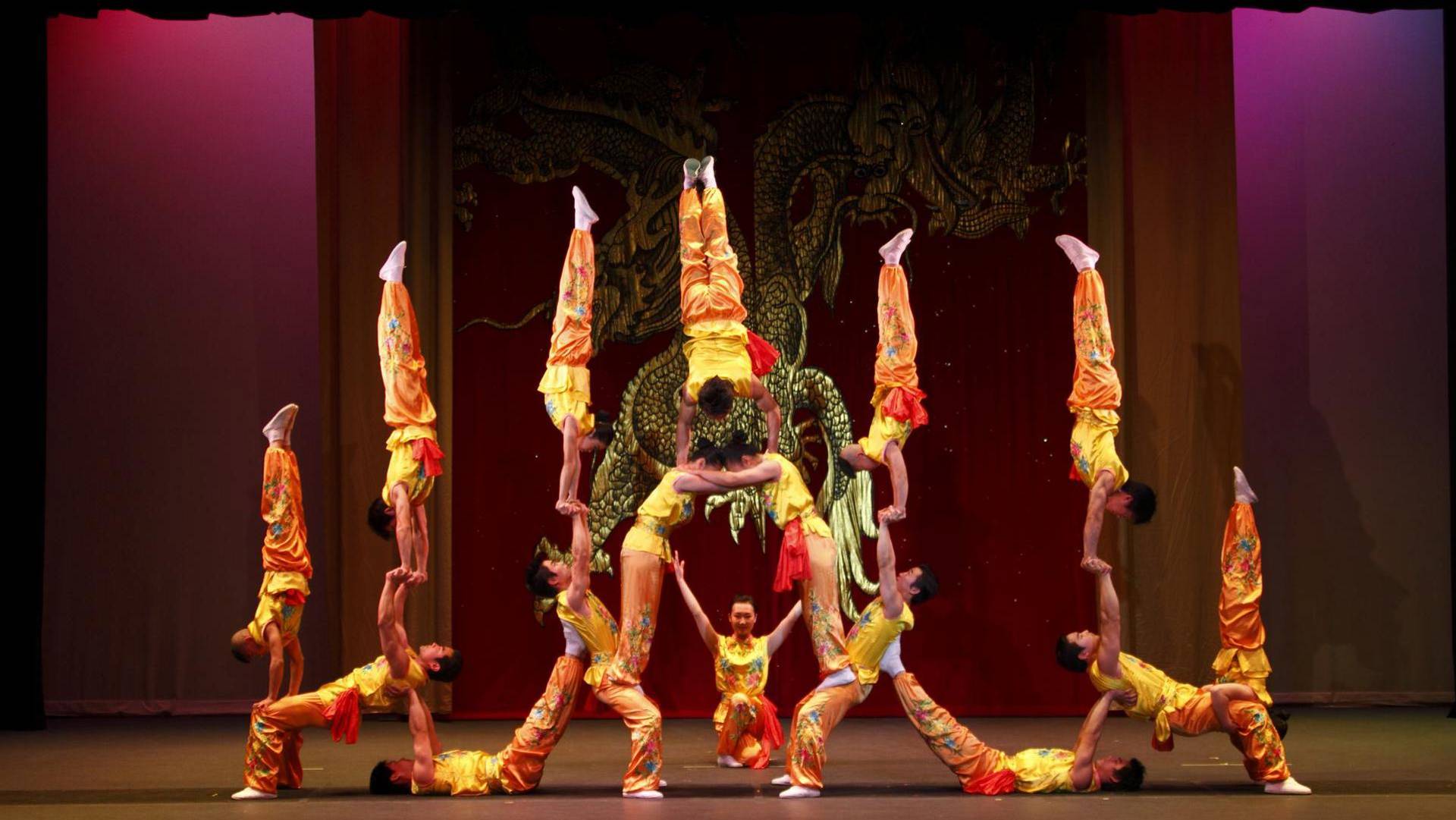 The Peking Acrobats performing on stage. 16 acrobats in silky orange pants and yellow sleeveless tops form a human pyramid with handstands and lifts.