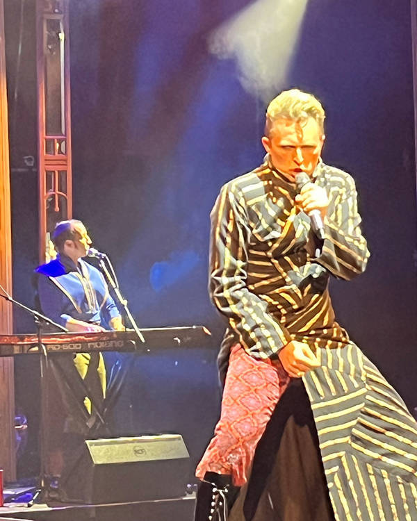 A man in a black and yellow striped longcoat with red pants sings on a stage.
