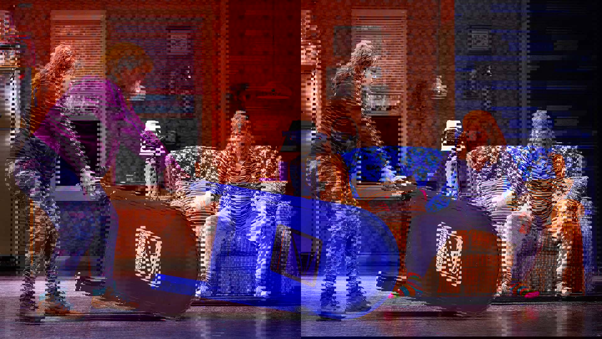 Two women with light complexions have an animated discussion while woman on left drags a blue US Postal Service mailbox across the ground.