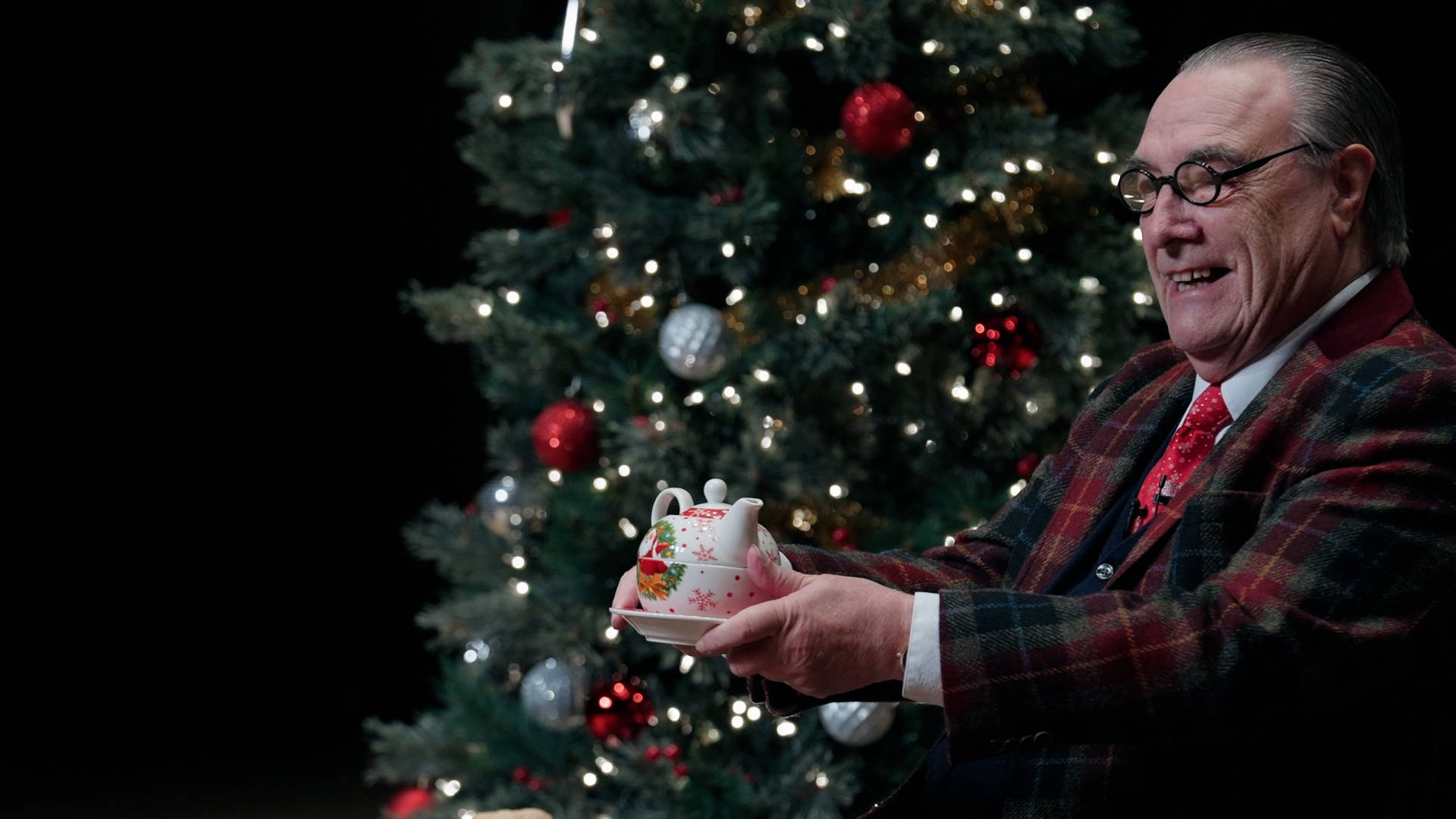 An older, white man with a receding hairline and black glasses in a blue suit and plaid jacket. He is sitting in front of a Christmas tree holding a teapot.