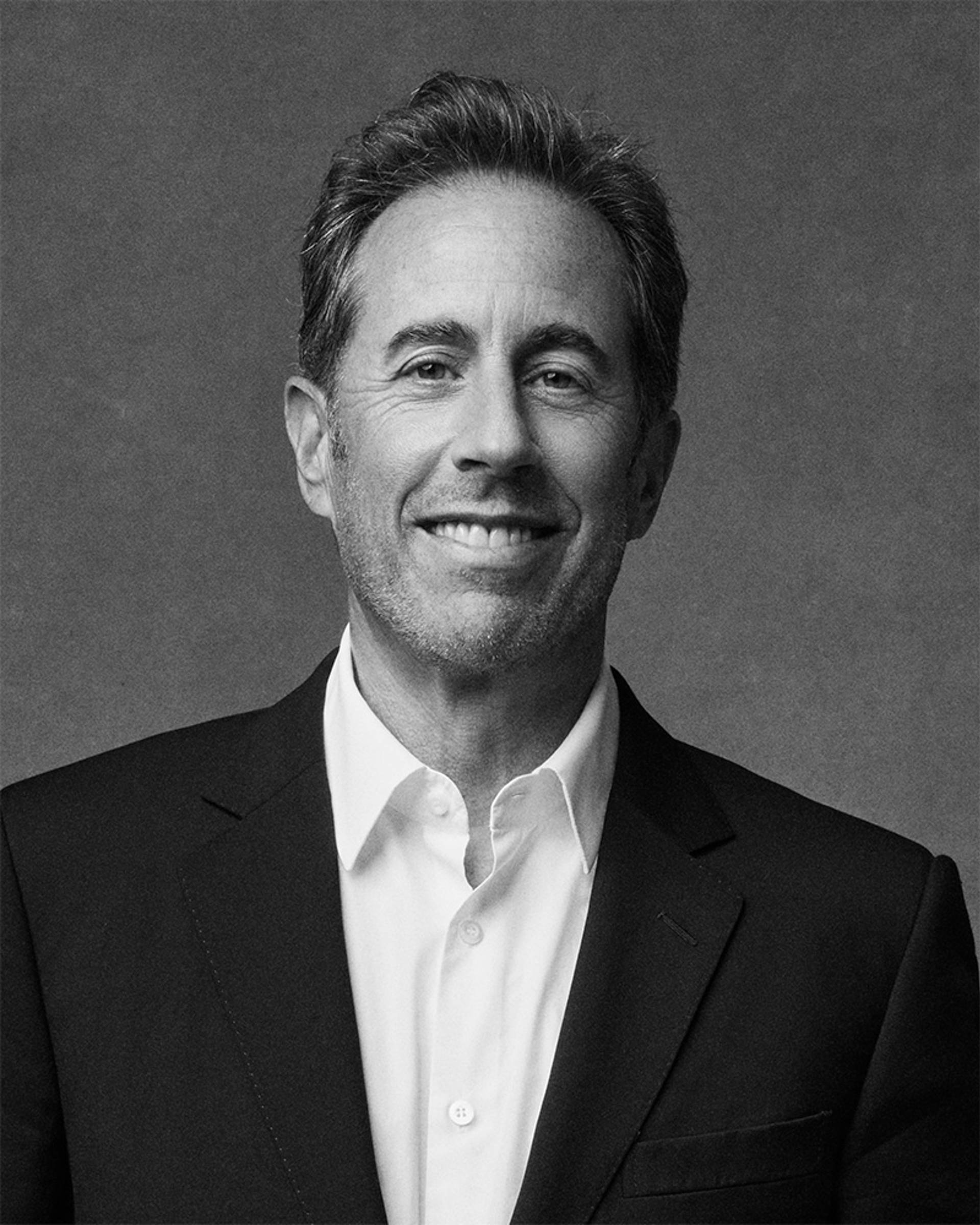 A greyscale image of Jerry Seinfeld, a middle aged man with light complexion, short dark hair, and a black suit with no tie. 