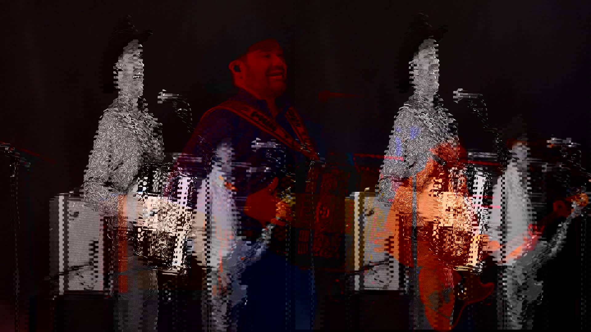 A hispanic man in a black cowboy hat, blue paisley collared shirt and jeans playing the accordion on stage.