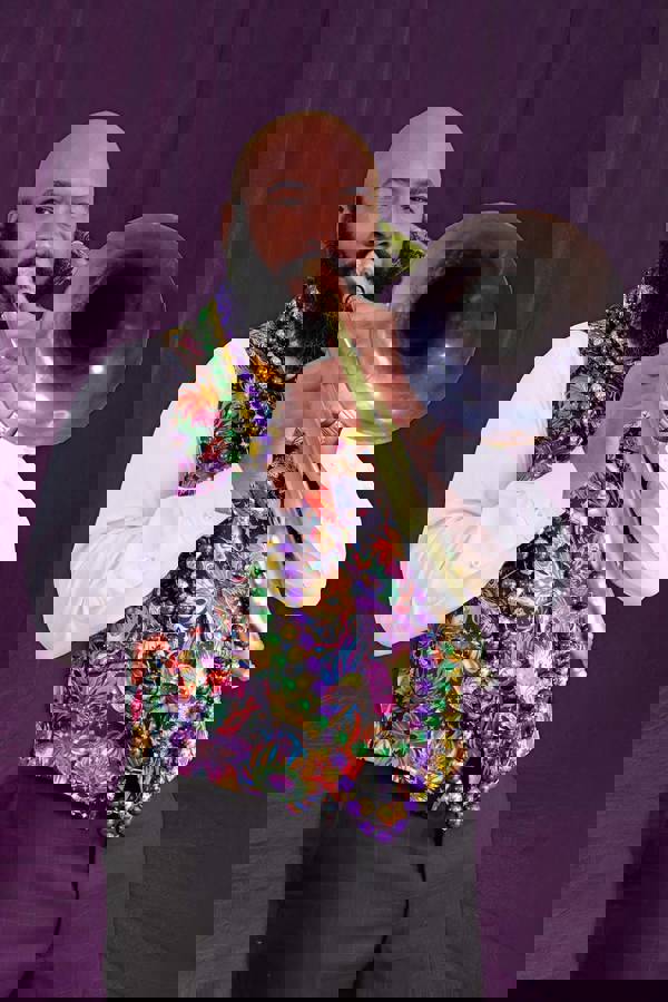 A bald black man with a large beard playing the trombone. He is wearing a black suit, no jacket, with a vibrant Mardi Gras designed vest and colorful bead necklaces.