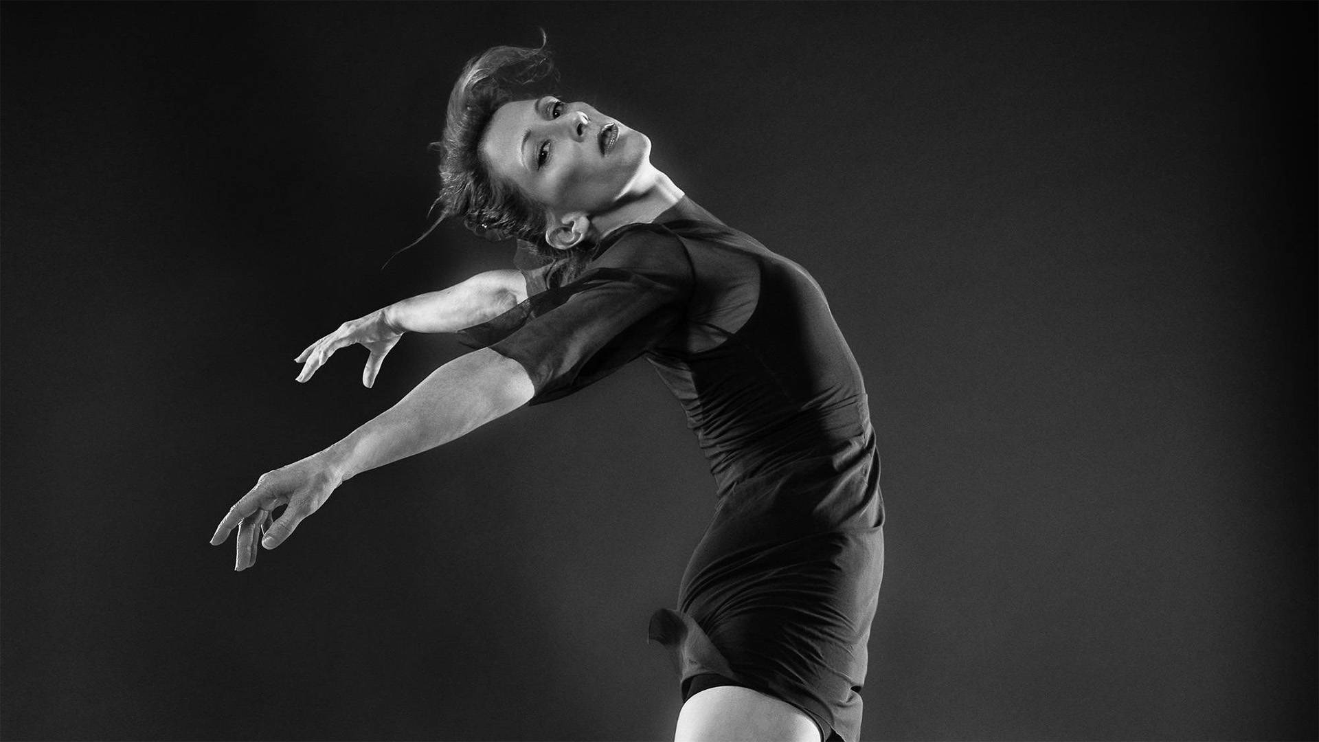 Black and white photo of a dancer in a black outfit with opaque material. Her arms are extended backwards.