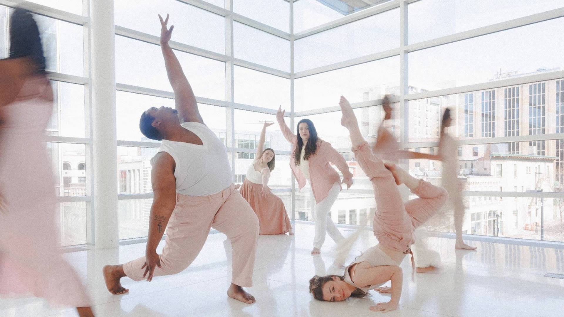 Dancers in various flowing light pink and white outfits striking dramatic dance poses against large windows looking out on buildings in the daytime.