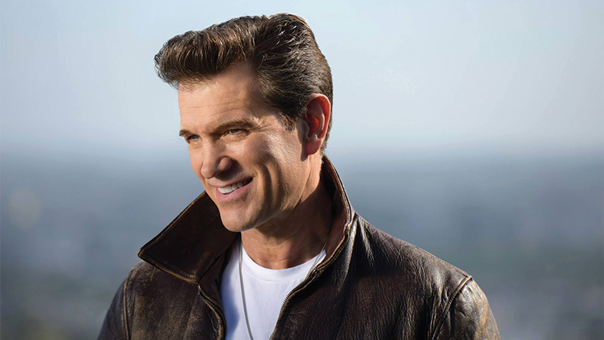 Chris Isaak smiles and slightly squints. He has coifed short brown hair and is wearing a beat leather jacket and white tshirt