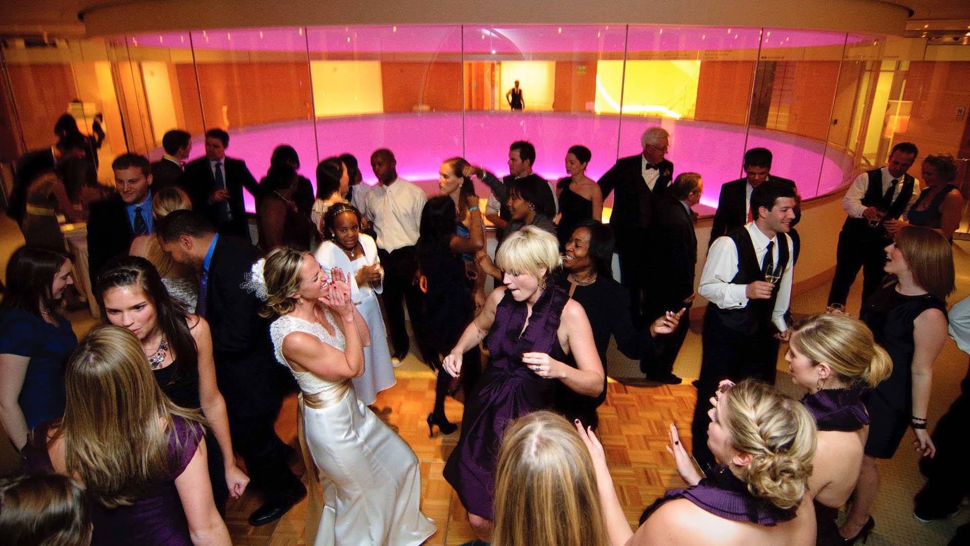 A bride dancing with friends on a dance floor. There is a circular glass window behind them with pink lights.