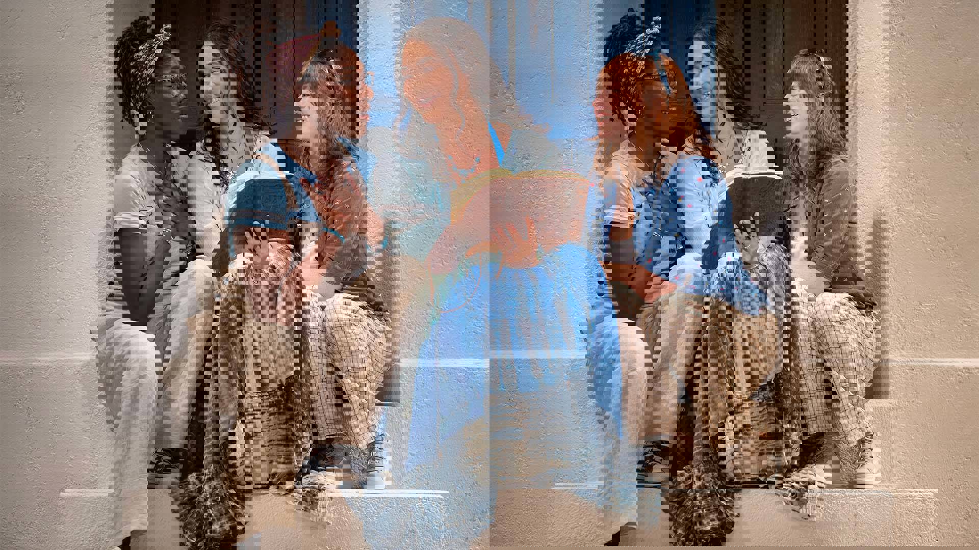 Three young women in blue and tan outfits sitting on steps looking at a book the center person is holding.