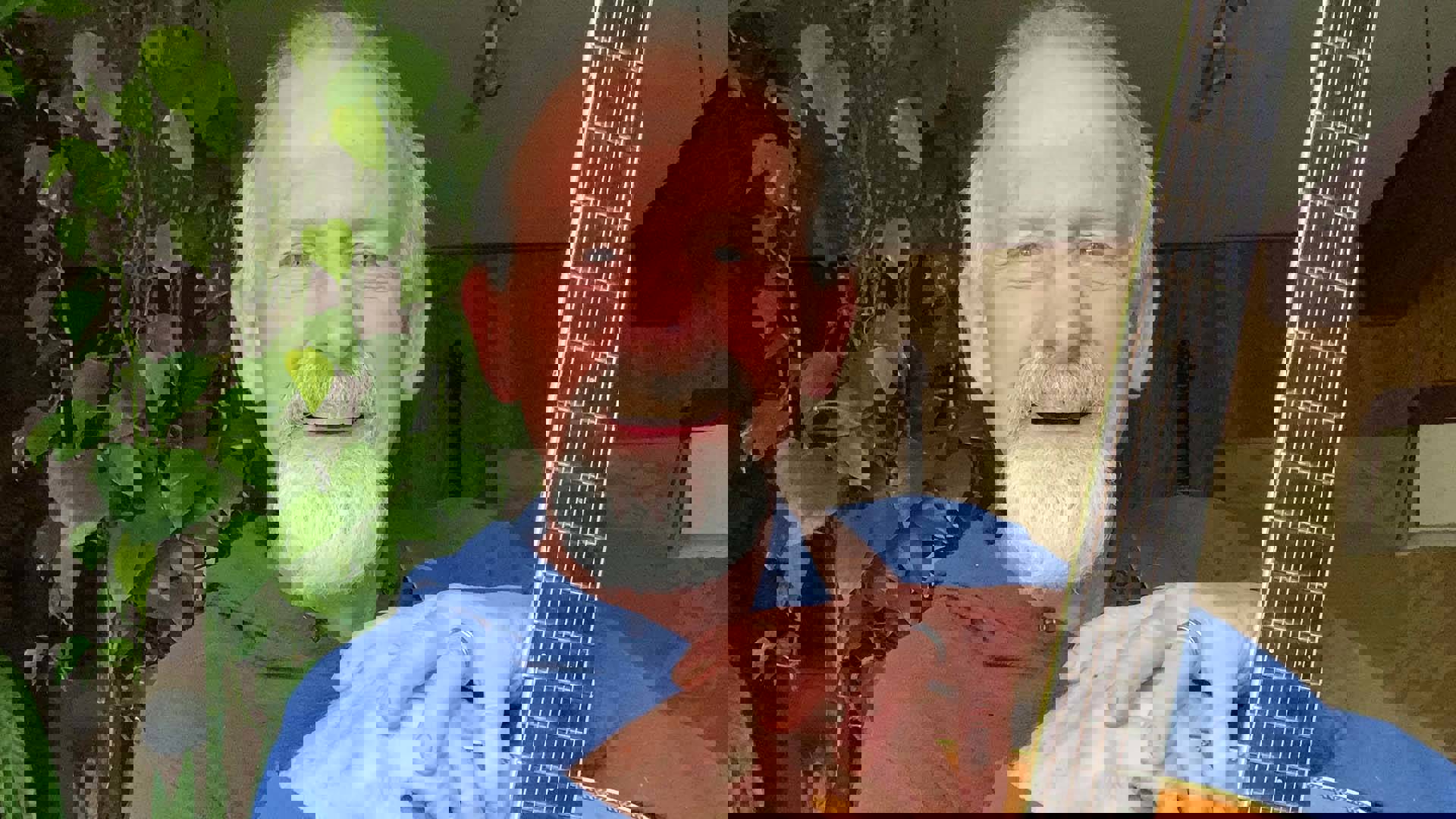 A Caucasian man with white hair and a goatee wearing a blue hoody smiling with a guitar on his lap.