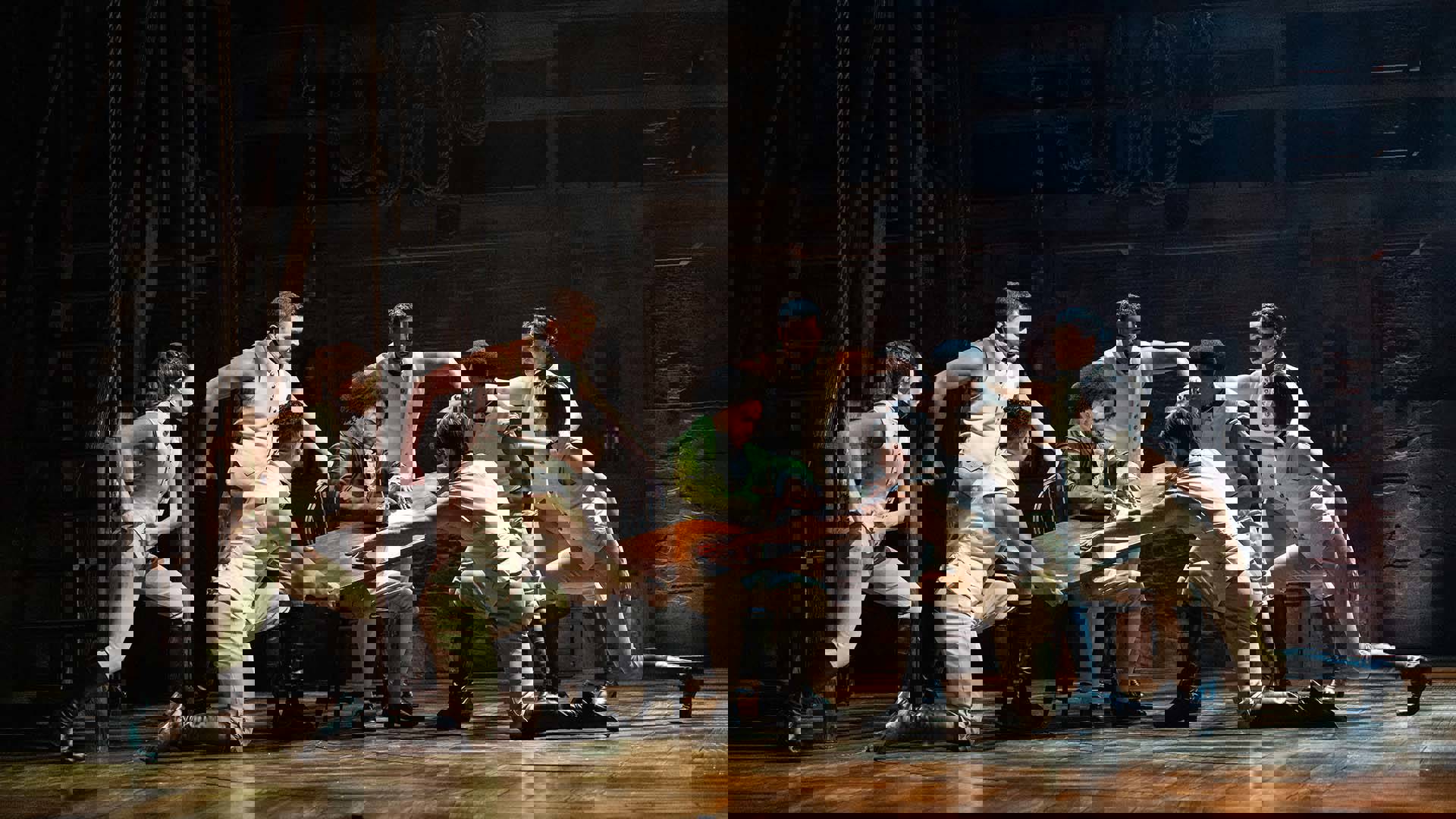 A group of men in beige uniforms hold a floating desktop while a man with dark complexion in a green jacket sits and writes on the desk.