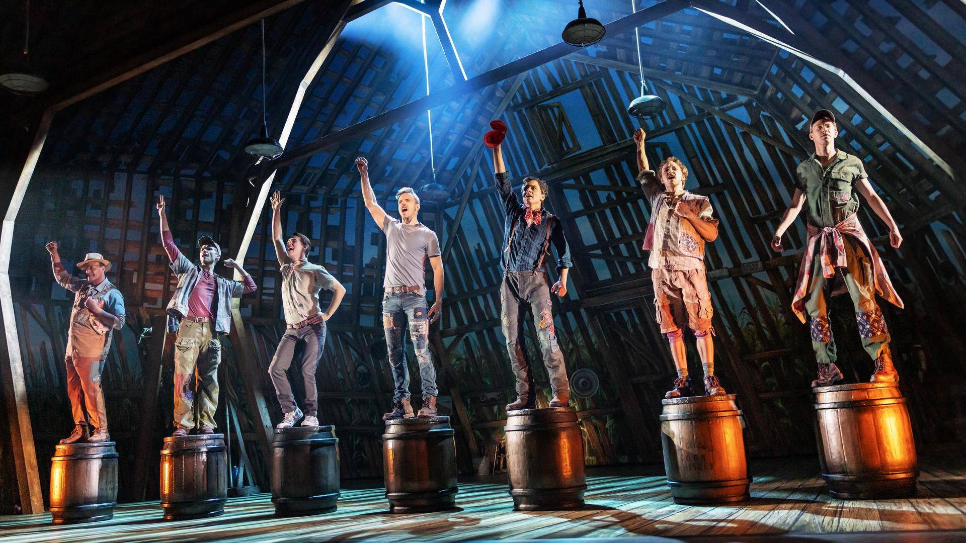Seven people stand on top of whiskey barrels in the barn. They have lights over them. They are all singing and have one hand in the air.