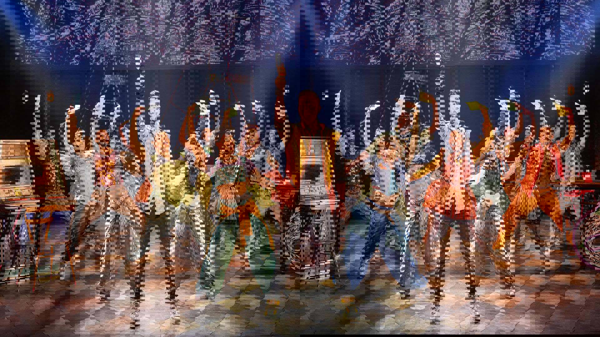 A man with a yellow and brown vest with light complexion holding a quill into the air while an ensemble behind him strikes similar poses holding pieces of paper into the air.