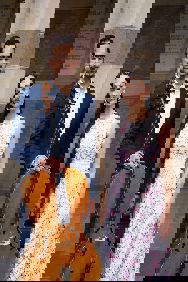 A man with medium complexion, dark hair and a light beard in a blue suit coat propping up a cello. To his left is a young woman with medium complexion, long brown hair, and a white dress.