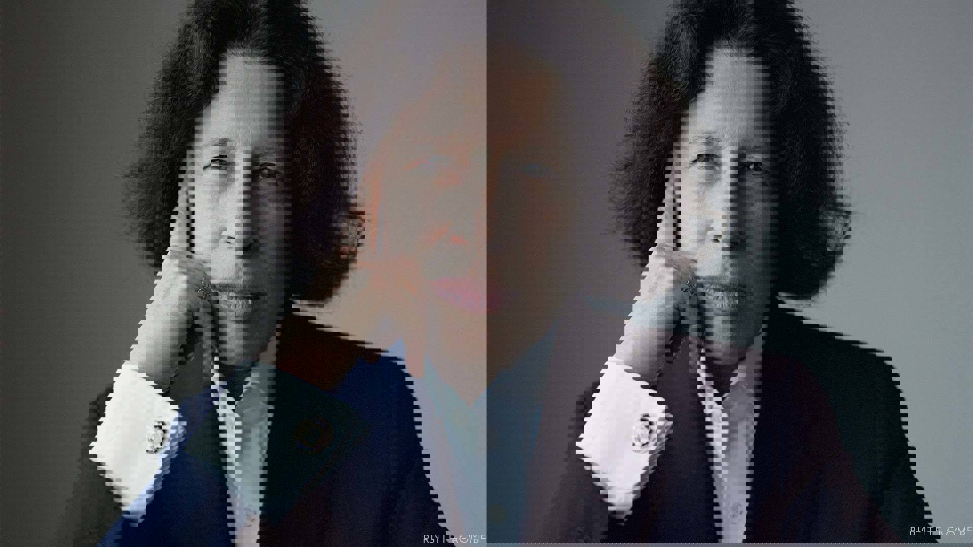 Headshot of Fran Lebowitz. She has medium length dark hair, dark eyes and is resting her head against her hand. She is wearing a blue suit and white buttonup shirt.