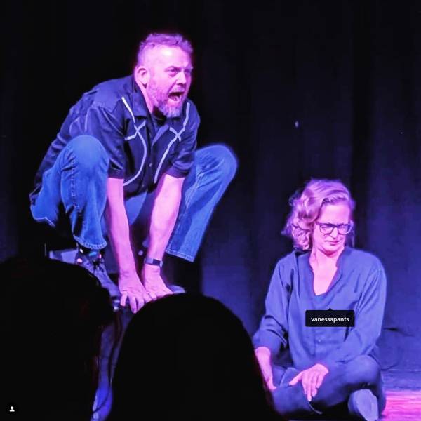 Jess Schuknecht performing improv. He is a white man with short hair and a beard. He is squatting on a stool while another performing with light complexion and medium blond hair makes a funny squinty face