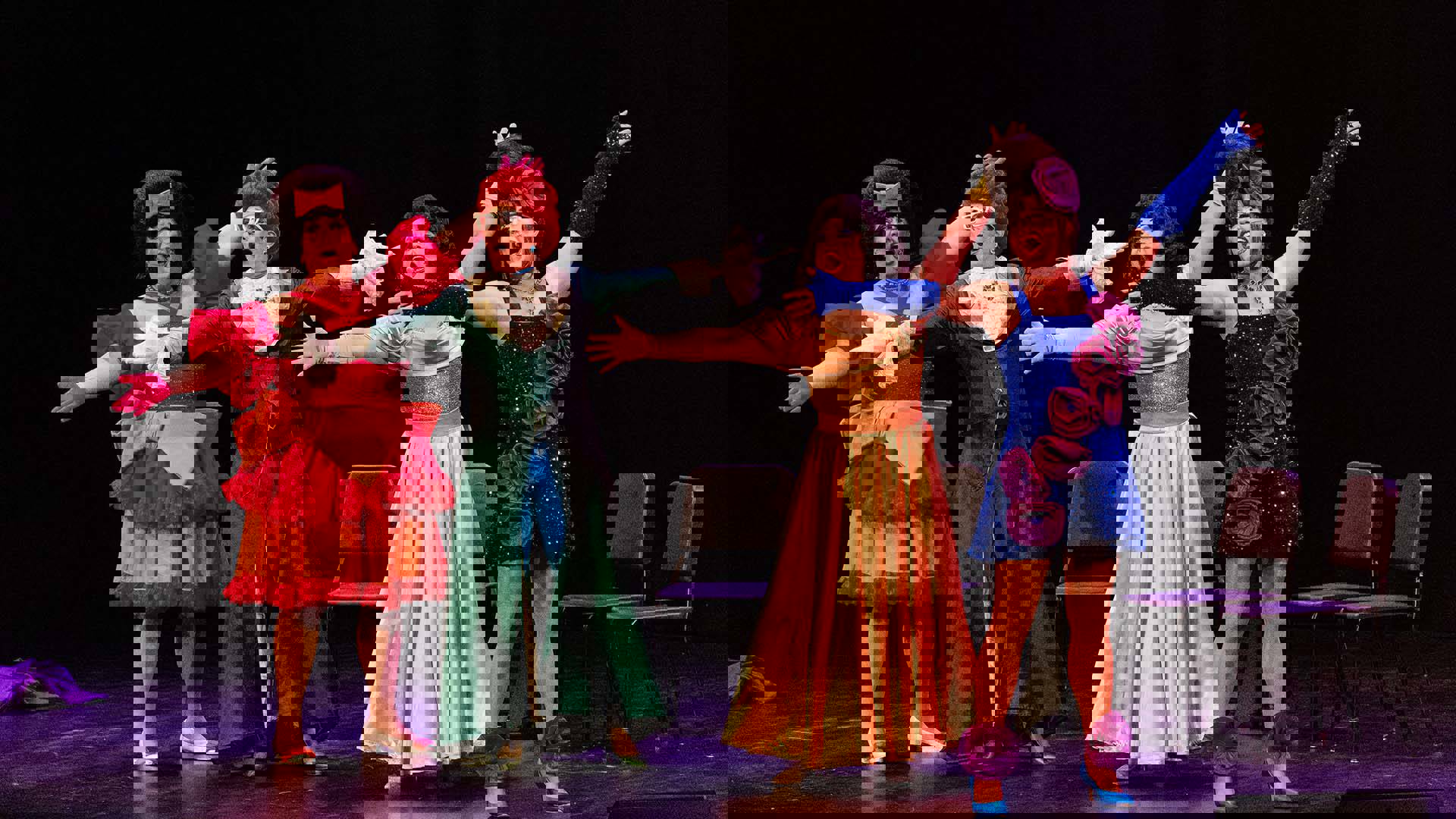 Four performers in colorful costumes and large wigs sing while holding their arms outstretched.