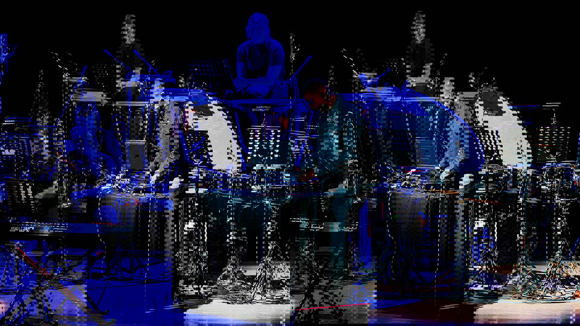 A man with dark complexion and stylish dark clothing DJing on a stage. with orchestral equipment behind him.