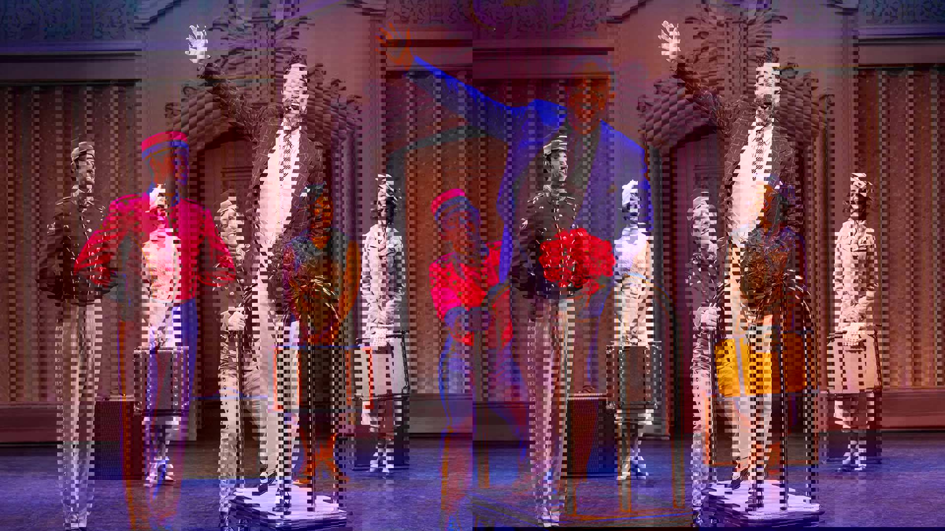 A man with light complexion, dark short hair and a dark, greying beard, wearing a white suit with a blue suit jacket and oxford shoes. He stands on a hotel luggage cart holding a bouquet of red roses, smiling with the other hand out wide. Two bellboys in red costumes stand behind hime smiling.