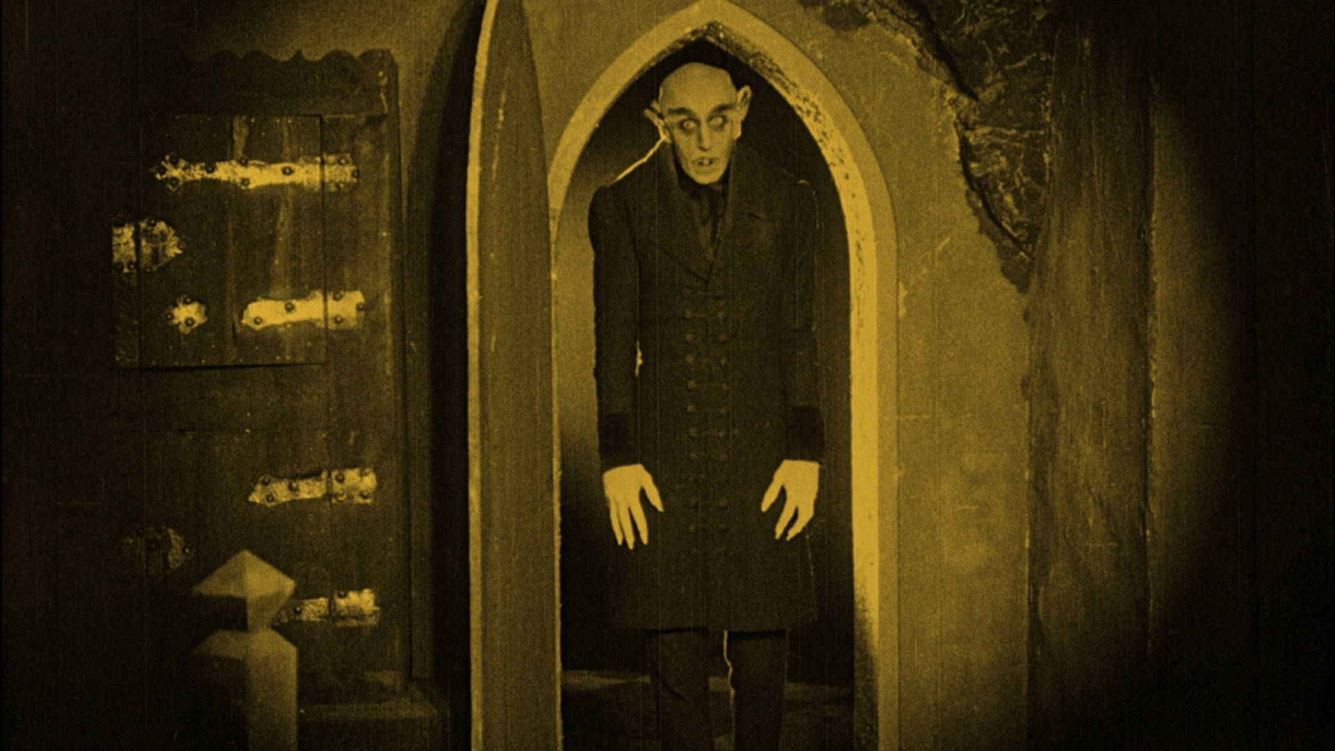 Vintage monotone image of a tall scary figure with long fingers, a bald head, and a dark outfit on standing in a doorway and looking at the viewer.