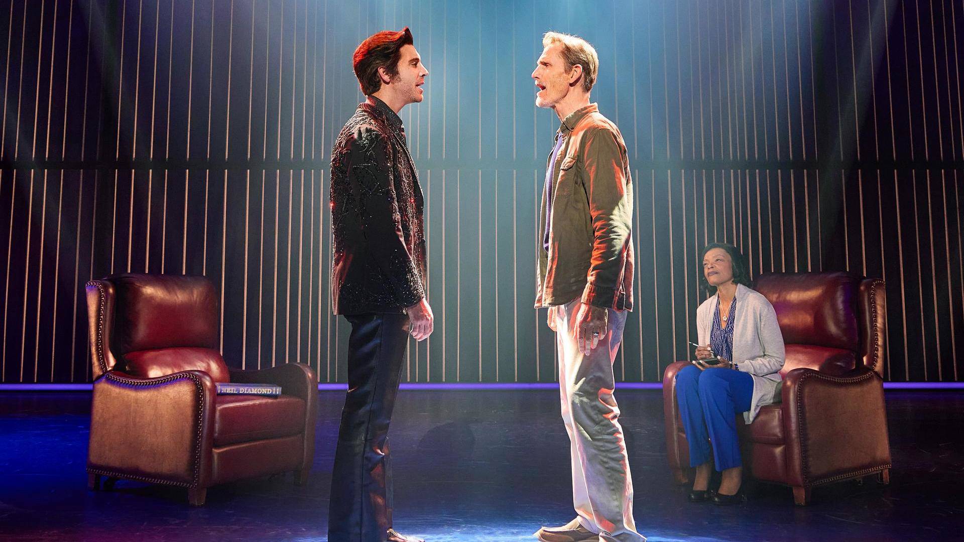 Two men with light complexion and short hair face eachother and are singing. A woman in the background is sitting on a leather seat and listening.