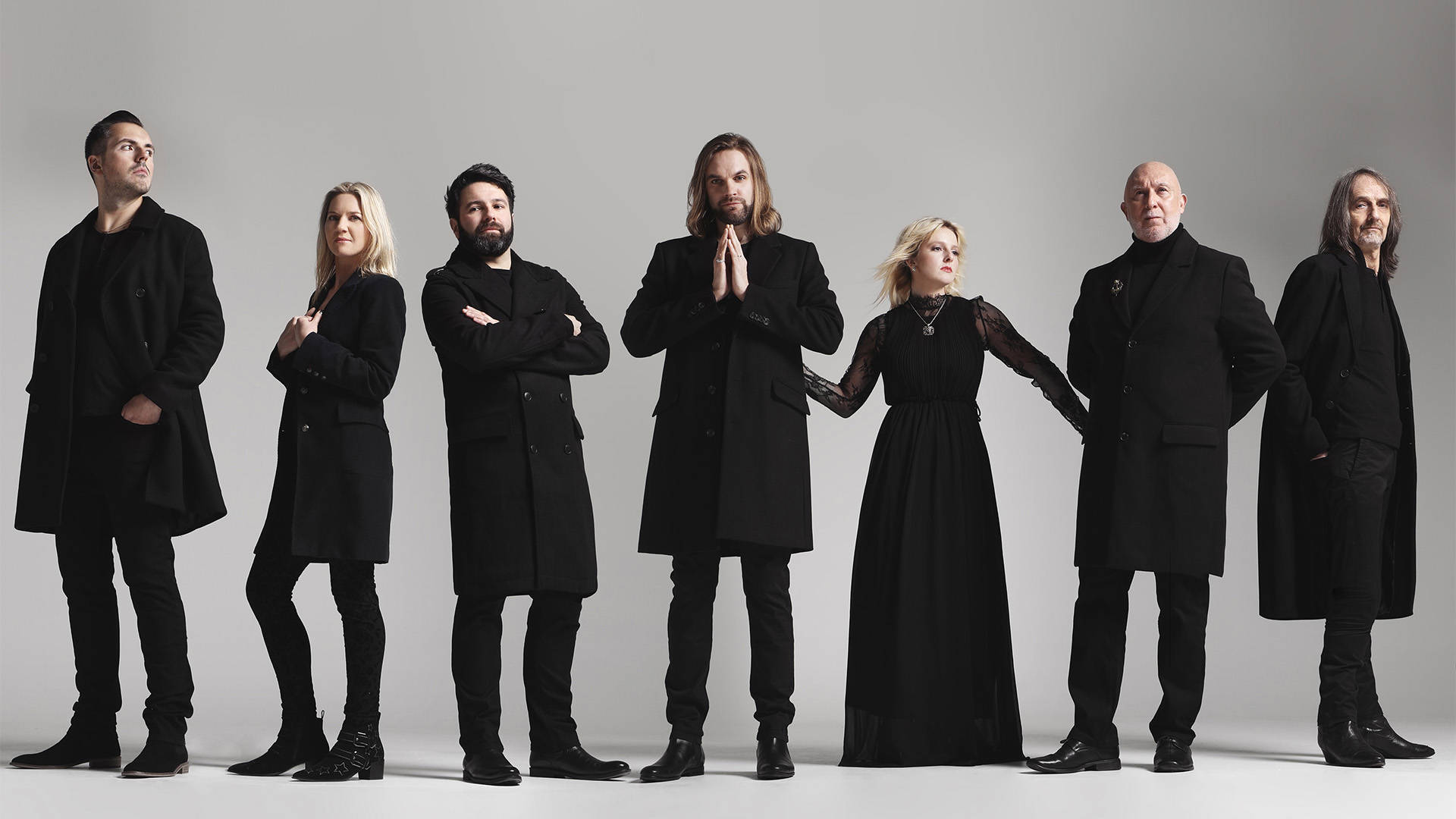 "Rumours of Fleetwood Mac" band: seven caucasian people standing in all black against a grey backdrop. 