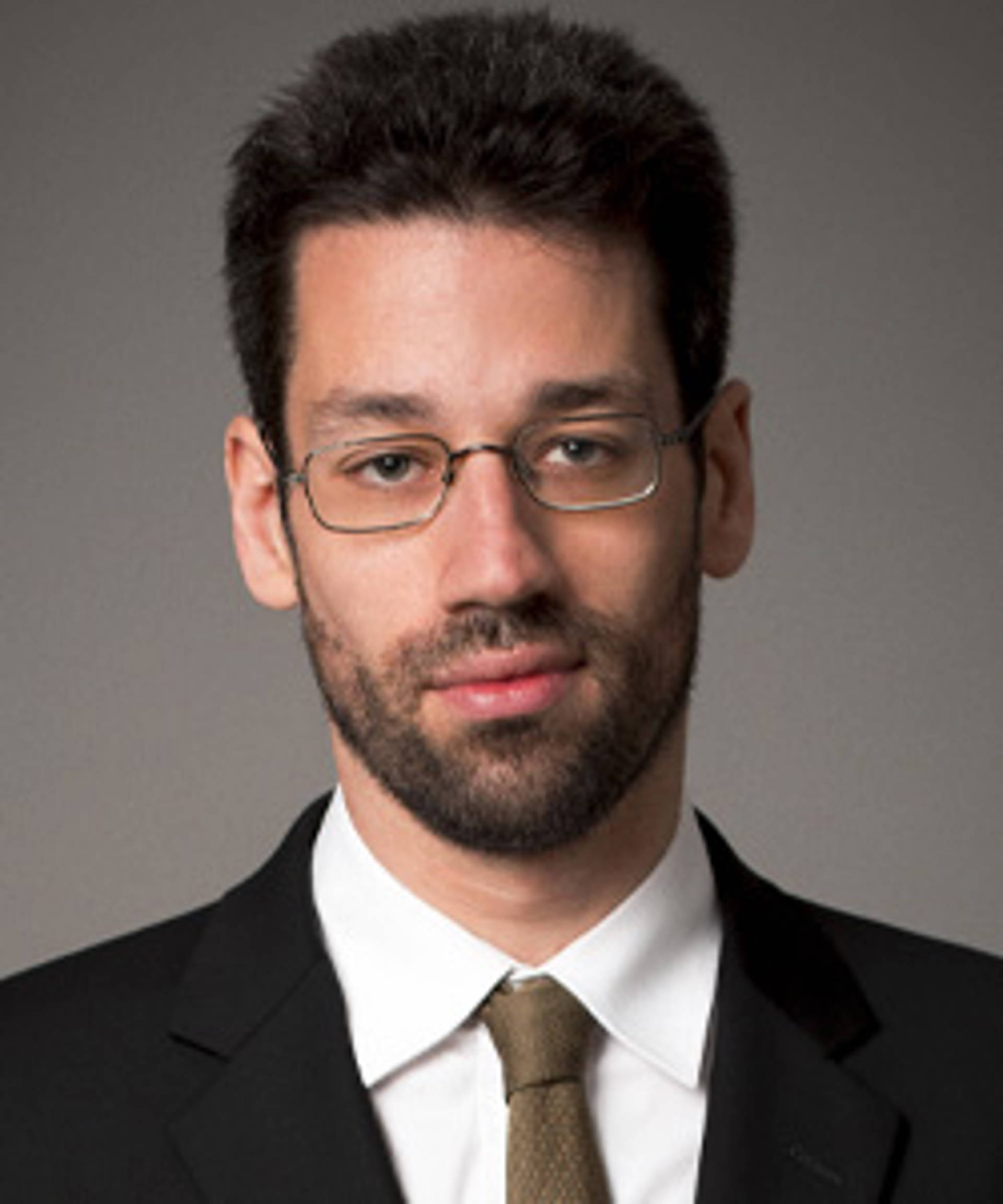 A young man with light skin, a short dark beard, glasses, and dark hair.